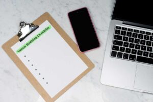 Clipboard titled “Venue Booking Checklist” next to a smartphone and laptop on a marble desk, representing digital tools for booking shows and emailing venues.