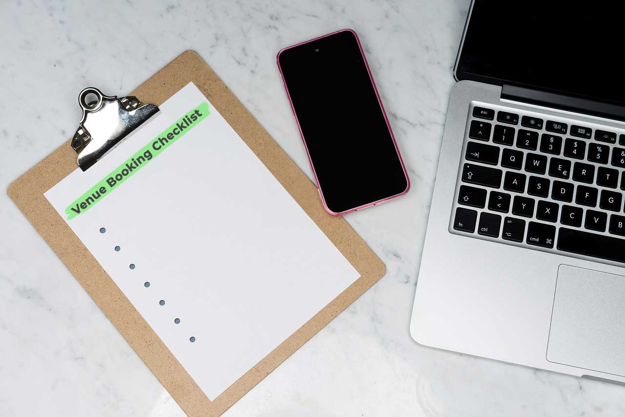 Clipboard titled “Venue Booking Checklist” next to a smartphone and laptop on a marble desk, representing digital tools for booking shows and emailing venues.